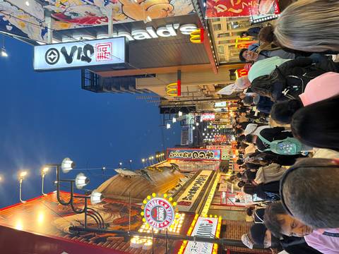       Busy pedestrian street in Osaka at dusk lined with bright restaurant signs and fast-food icons.
  