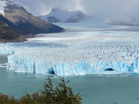       Massive blue wall of Perito Moreno Glacier meets turquoise lake water with distant mountains looming.
  