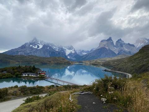       Turquoise lake reflecting snow-capped mountains and the iconic Cuernos del Paine with a small lodge on a narrow causeway.
  