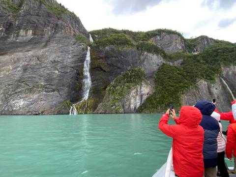       Bundled tourists in red jackets photograph tall waterfalls cascading down sheer rock faces into a turquoise fjord.
  