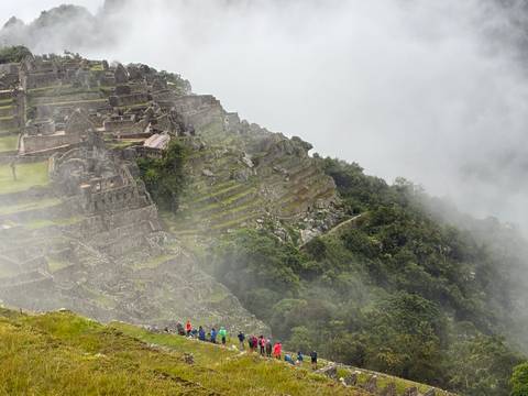       Oblique view of Machu Picchu’s steep terraces and stone buildings descending into lush jungle with tiny hikers below.
  