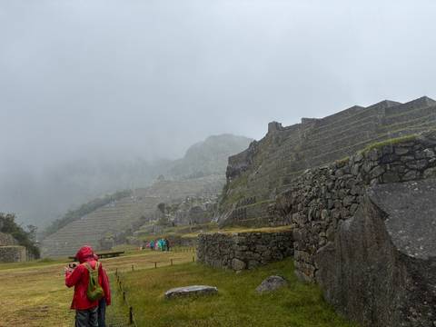       Rain-soaked visitors explore lower terraces of Machu Picchu on a foggy day, stone walls darkened by moisture.
  