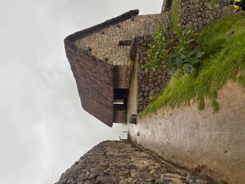       Stone hut with thatched roof sits on a misty path lined by ancient walls at Machu Picchu.
  