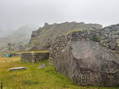       Stone terraces and ancient stone walls of Machu Picchu shrouded in mist, with small groups of tourists in colorful ponchos on the grassy foreground.
  