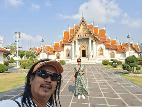       Selfie of two travelers in front of ornate Bangkok temple with orange roofs and manicured garden.
  