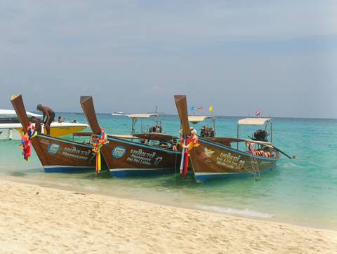      Three traditional long-tail boats with colorful garlands rest in shallow turquoise water along a tropical beach.
  