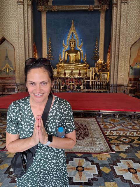       A woman performs a respectful wai gesture inside an ornate Thai temple with a golden Buddha altar.
  