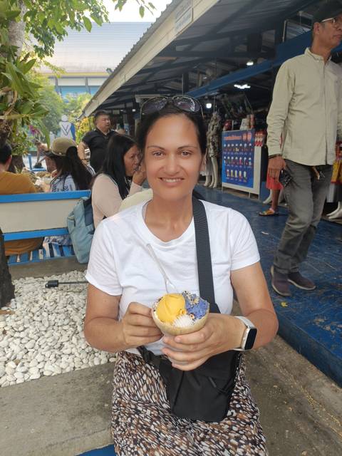       Smiling woman holds a colorful cup of ice cream at a busy outdoor market stall.
  