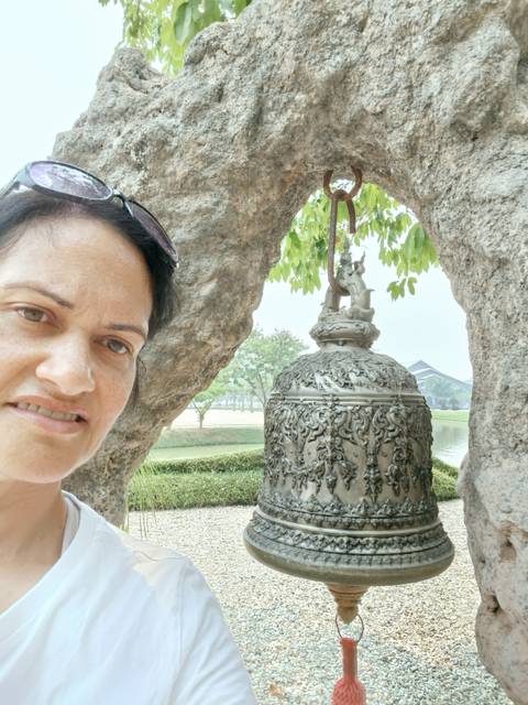       Selfie beside an ornate metal bell hanging from a tree with landscaped gardens in the background.
  