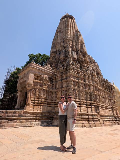       Smiling couple posing in front of an ornate sandstone temple covered in intricate carvings under a clear sky
  