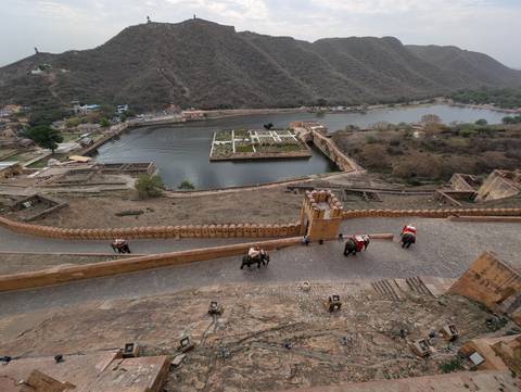      High view over Amber Fort ramparts, elephants with riders climbing the cobbled path beside a lake and gardens
  