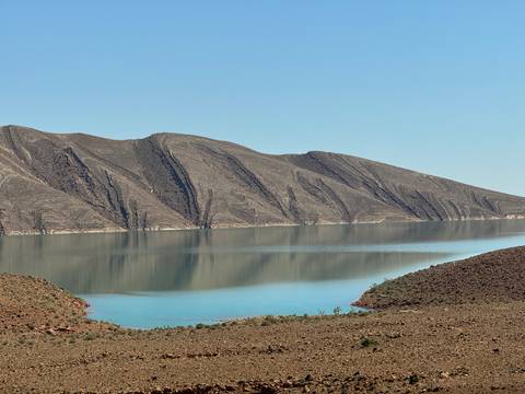       Still blue reservoir mirroring striated brown mountain slopes on a clear day
  