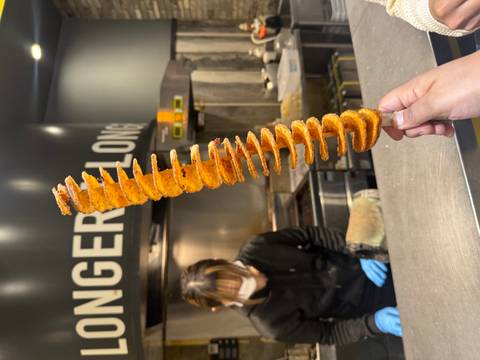       Hand holding a spiral fried potato snack while a masked vendor prepares food at a stall.
  