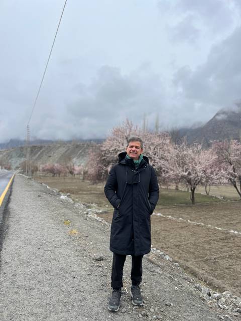       Man in dark coat standing roadside with blooming pink trees and misty mountains behind
  