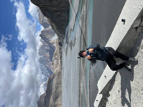      Traveler sits on ledge taking photo with braided riverbed and jagged mountains behind
  