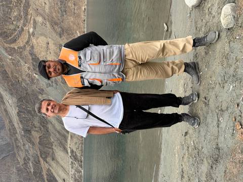       Two men posing on a lakeshore with steep rocky mountains rising behind the calm water.
  