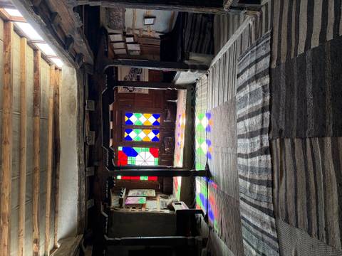       Dimly lit traditional room with timber beams and multicoloured stained-glass windows casting light onto woven rugs.
  