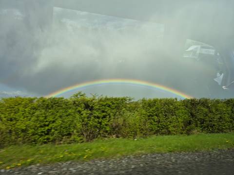       Vibrant rainbow arcs above a lush roadside hedge below brooding grey clouds, viewed through a vehicle window.
  