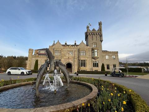      A grand stone castle hotel with a decorative fish fountain in front and an Irish flag flying from the tower on a clear day.
  