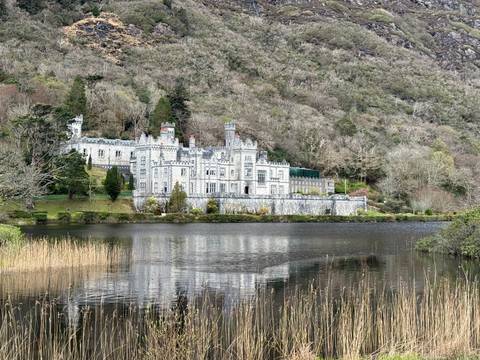       Kylemore Abbey, a grey neo-gothic castle reflected in a calm lake and backed by forested mountains.
  