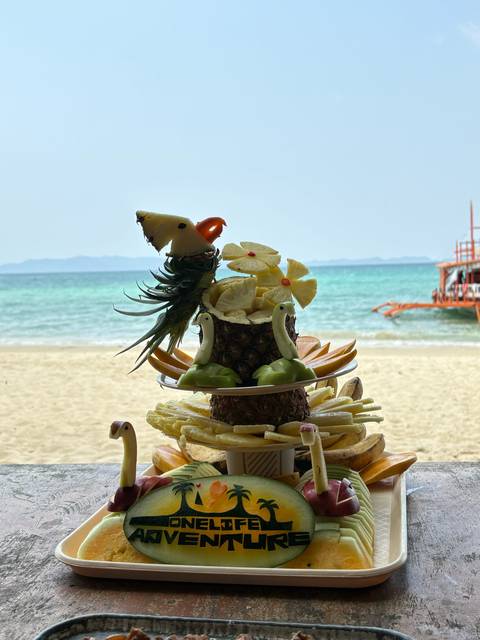       Elaborate tropical fruit carving shaped like birds and flowers set on a platter with white-sand beach and turquoise sea behind.
  