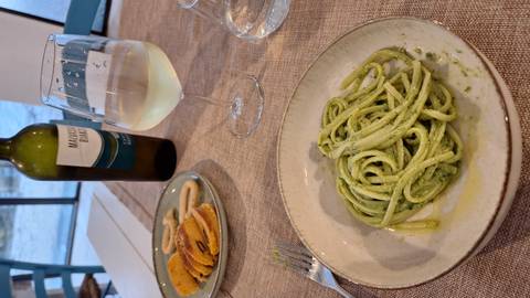       A bowl of pesto linguine with white wine, snacks and a bottle on a rustic table.
  