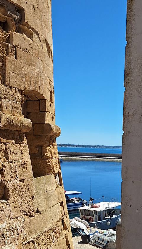       View between weathered stone bastions toward calm blue sea and distant shoreline.
  