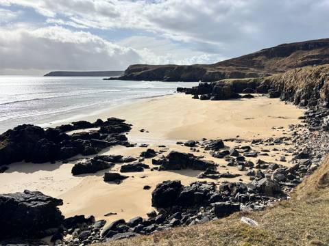      Secluded sandy beach with dark rocky outcrops and shimmering sea under partly cloudy skies
  