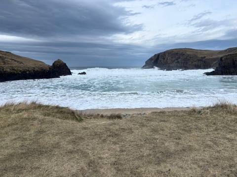       Wild waves crash into rocky sea stacks and cliffs beneath a moody grey sky on a remote Scottish bay
  