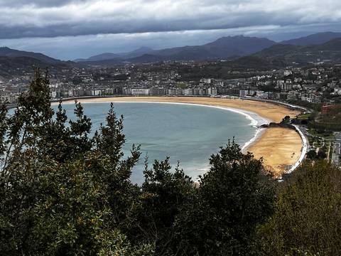       Panoramic view from hillside of a broad crescent-shaped urban beach backed by a coastal town.
  