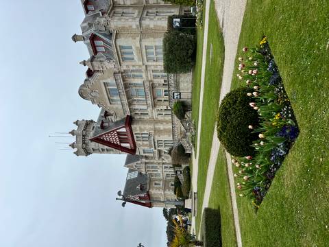       Elegant historic mansion with turrets and red timber detailing surrounded by formal lawns and flower beds.
  