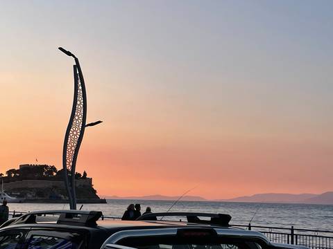       Silhouettes of people fishing beside the sea at sunset with an island fortress and modern sculptural streetlight.
  