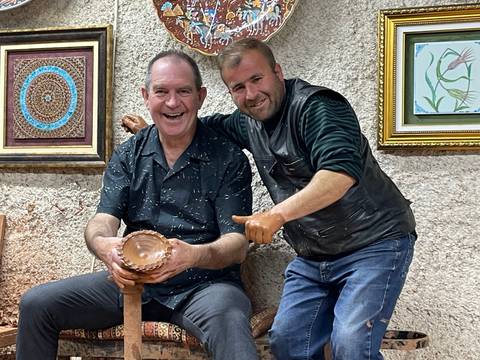       Two smiling men in a pottery workshop displaying a freshly shaped clay bowl, hands covered in clay.
  