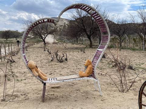       Heart-shaped metal bench in sandy, barren landscape inhabited by teddy bears amid leafless trees.
  
