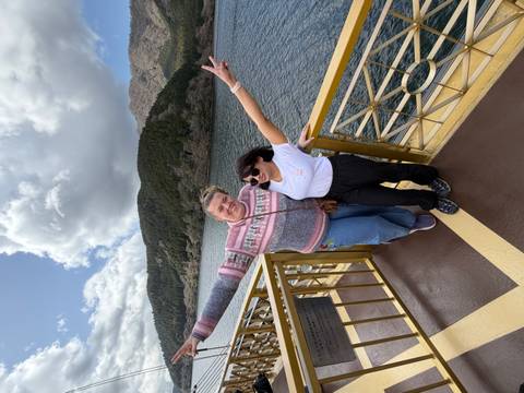       Two happy friends spread their arms on a lakeside ship’s deck with forested hills and water behind.
  
