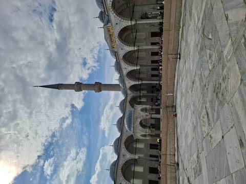       A grand mosque courtyard with elegant domes and a slender minaret against a partly cloudy sky.
  