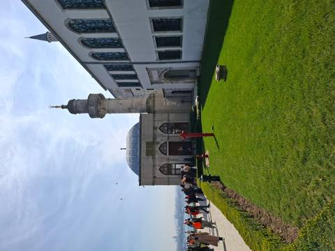       An exterior view of a small mosque with a green lawn and visitors milling about on a sunny day.
  