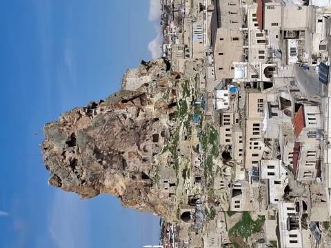       A towering rock citadel rises above a traditional stone town under clear blue skies.
  