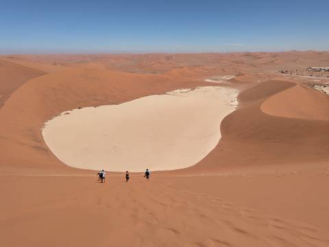       Tiny hikers descend massive orange sand dune into a pale clay pan amid endless Namib Desert waves.
  