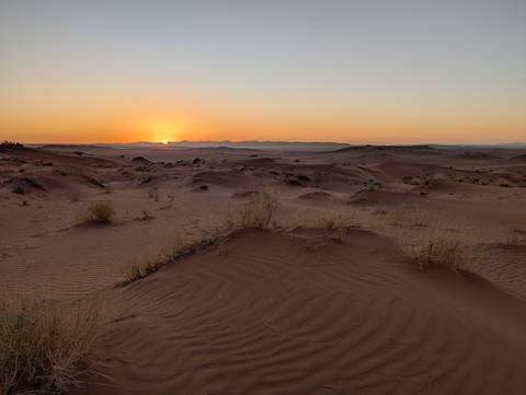       Soft orange sun sets behind distant mountains casting long shadows over rippled desert plains.
  