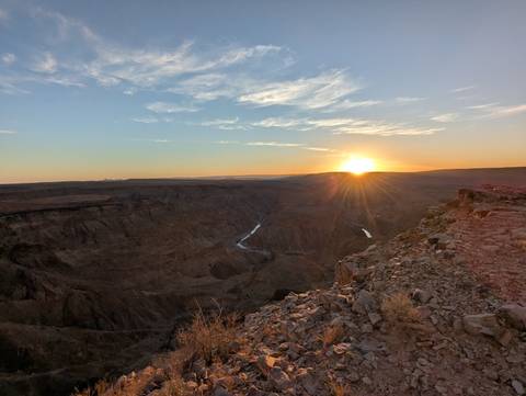       A dramatic view over a vast rocky canyon with a river far below, as the sun sets on the horizon under a clear sky.
  