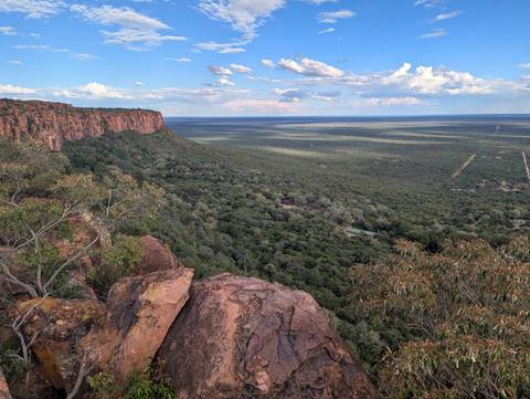       A lush green forest stretches to the horizon viewed from a rocky cliff edge under scattered clouds.
  