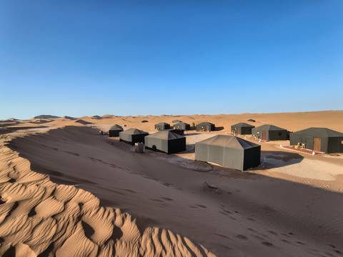       A line of black desert tents sits amid golden sand dunes beneath a bright blue sky.
  