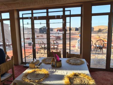       View from an indoor dining area looking out to desert camp fire pits and tents at dawn.
  