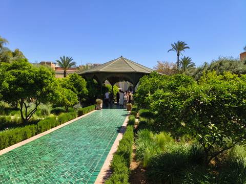       Visitors stroll along a turquoise tile pathway through lush green gardens toward a shaded pavilion.
  