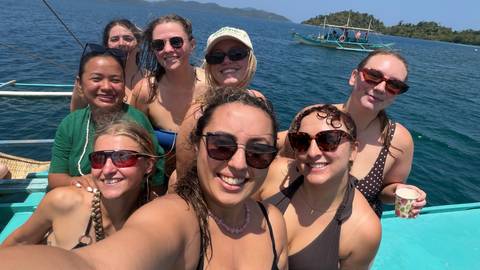       A smiling group of friends pose for a selfie on a boat in clear blue tropical waters.
  