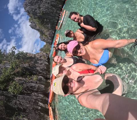       Friends stand waist-deep in crystal-clear lagoon water surrounded by towering limestone cliffs.
  
