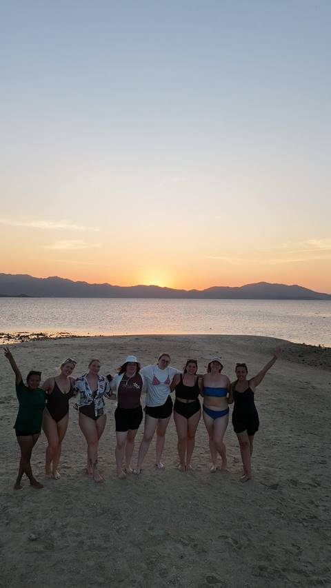       A group celebrates on a sandy spit as the sun sets behind distant mountains and calm sea.
  