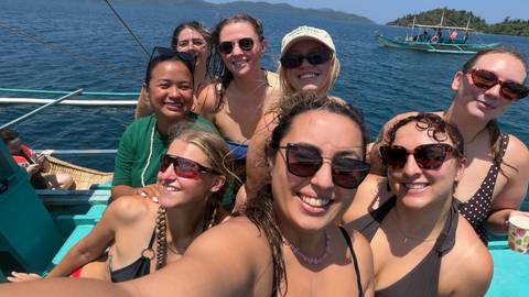       A happy boat selfie with turquoise water and an outrigger canoe in the background.
  