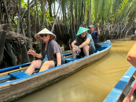       Tourists wearing conical hats paddle through narrow, palm-fringed canal in a wooden canoe
  